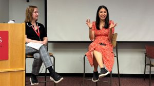 Two people are seated in high chairs at the front of a room. One is smiling and gesturing.