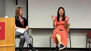 Two people are seated in high chairs at the front of a room. One is gesturing and smiling while the other looks on.