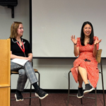 Two people sit on high chairs at the front of the room. One is smiling and gesturing her arms.