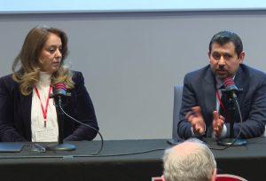 Two people sit at a table, with one gesturing their hands and speaking into a microphone