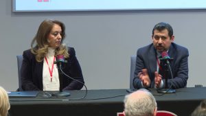 Two people are seated at a table. One watches as the other speaks into the microphone, hands gesturing.