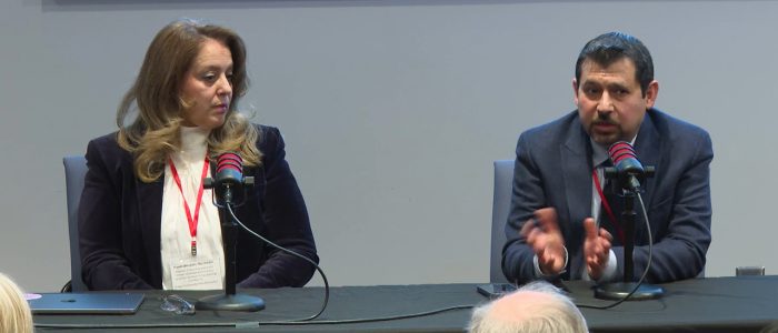 Two people are seated at a table. One watches as the other speaks into the microphone, hands gesturing.