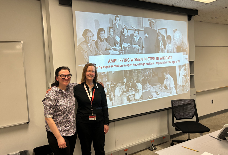 Two people pose smiling in front of a large screen with an image projected that reads, "AMPLIFYING WOMEN IN STEM IN WIKIDATA: Why representation in open knowledge matters - especially in the age of AI