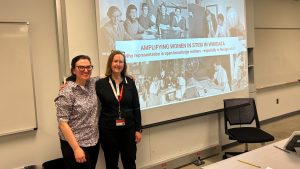 Two people smile standing in front of a large presentation screen that reads, AMPLIFYING WOMEN IN STEM IN WIKIDATA: Why Representation in Open Knowledge Matters - Especially in the Age of AI"