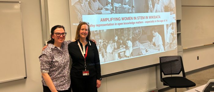 Two people smile standing in front of a large presentation screen that reads, AMPLIFYING WOMEN IN STEM IN WIKIDATA: Why Representation in Open Knowledge Matters - Especially in the Age of AI"