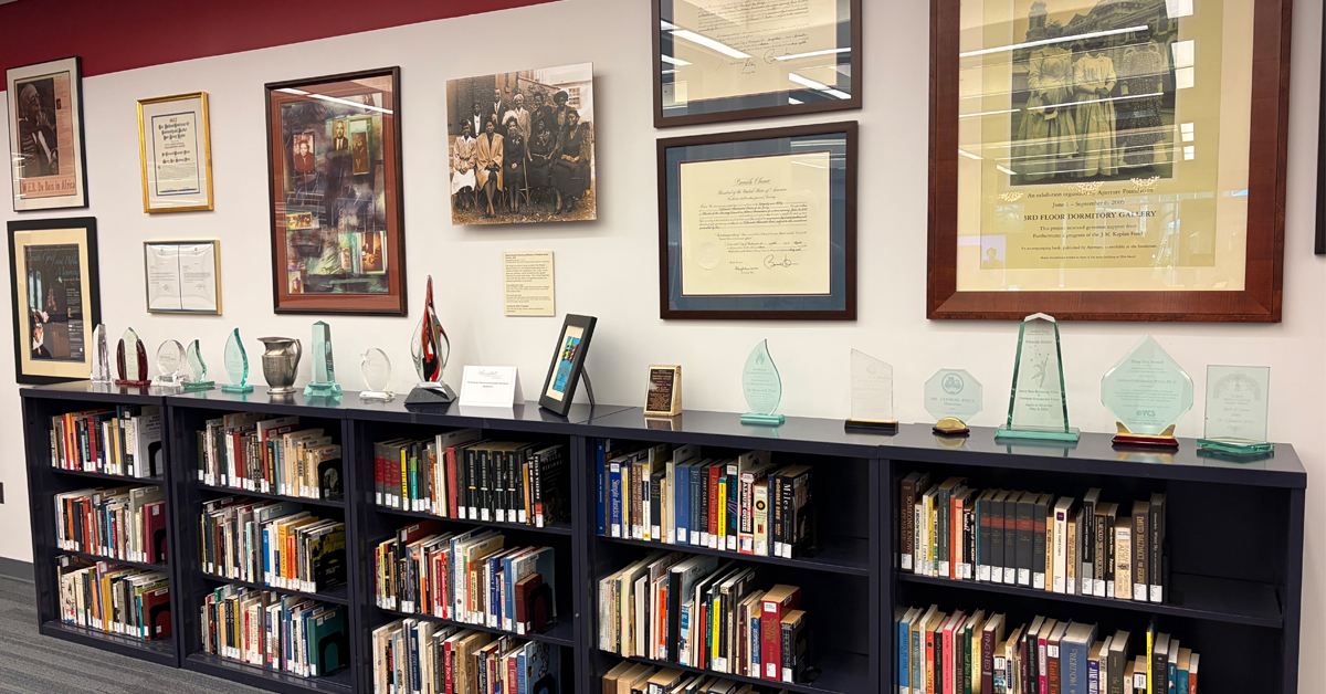 A long bookshelf against a wall with various artifacts displayed on top and tightly grouped framed images on the wall above.