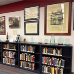 Full bookshelves lining a wall with artifacts placed on top and closely grouped framed pictures on the wall directly above.