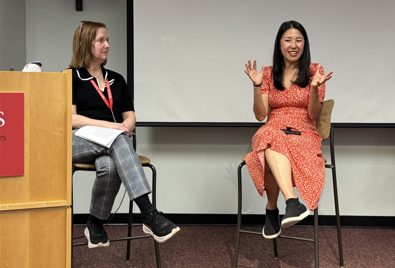 Two people are seated in high chairs at the front of a room. One smiles and gestures with her hands while talking.